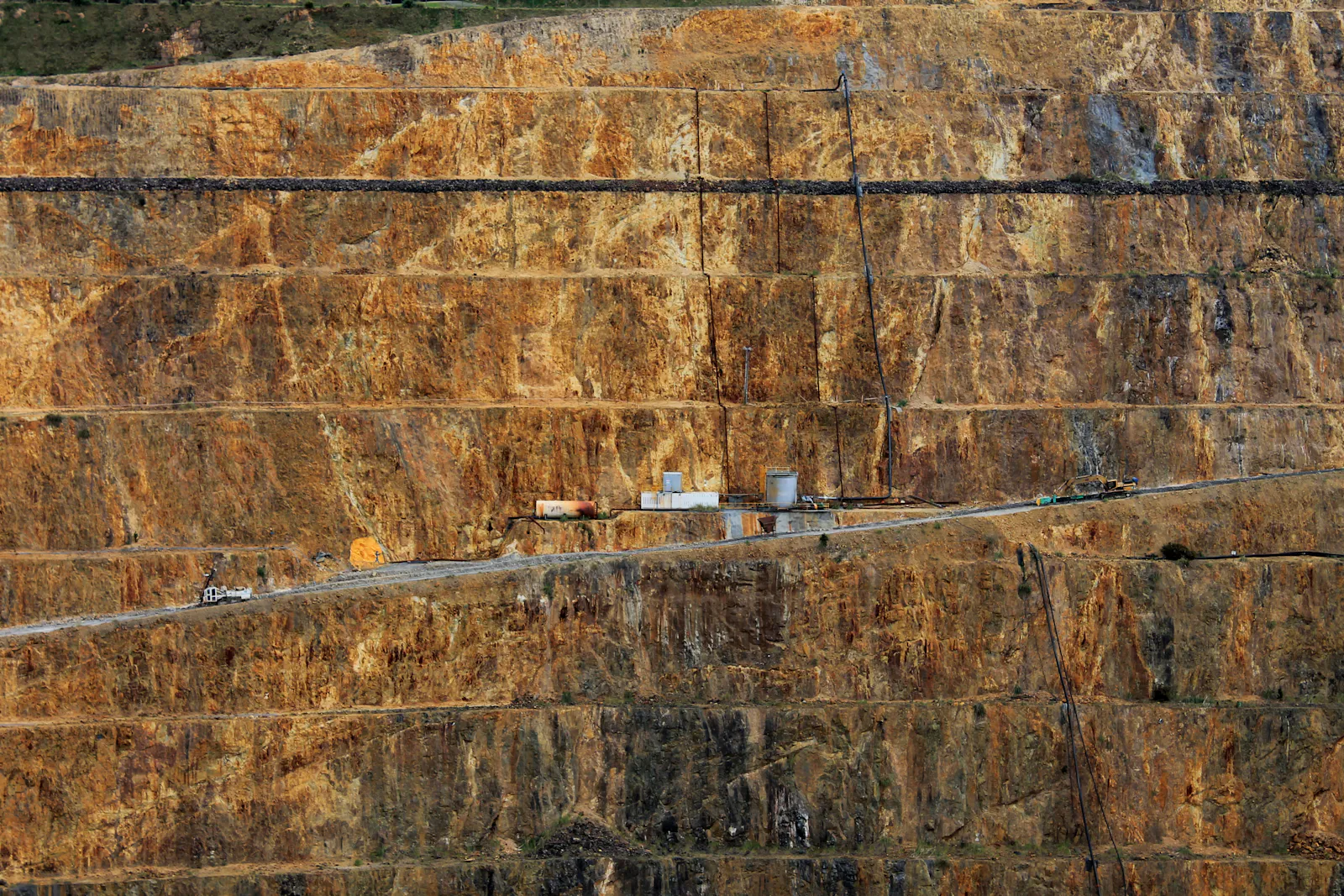 Terraced benches of an open-pit mine with a haul road and small buildings for scale